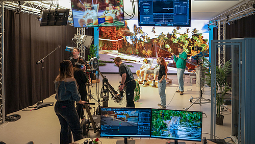 Nur fast wie im Urlaub: internationale studentische Filmproduktion im virtuellen Studio der Hochschule Mittweida. Junge Menschen im Studioraum mit Kameras, Scheinwerfen udn Monitoren. Im Bildhintergrund sitzen zwei Oersonen in Liegestühlen vor einer raumgroßen LED-Wand, die einen Palmenstrand zeigt.